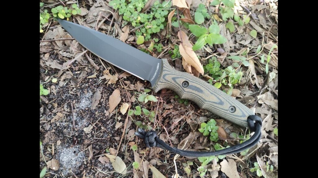 Fixed-blade knife with black blade and camo handle, lying on forest ground with lanyard attached.